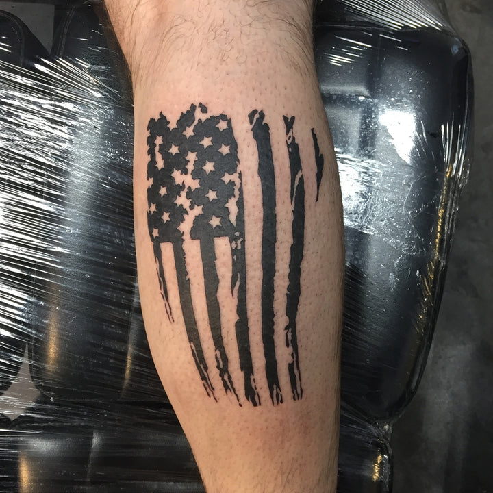 Tattoo of a torn american flag in black on the back of a mans calf.