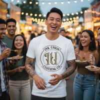 IU Black Badge Tee — Tattooed East Asian male model wearing white Ink Union Clothing Co. black badge brand art unisex graphic tee laughing with friends at outdoor street food festival with colorful stalls and string lights in background — alternative streetwear, tattoo artist t-shirt, unisex graphic tee, edgy cool fashion