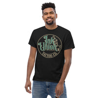 An attractive man wearing a black t-shirt with a gold circle containing fancy lettering in green and gold that says Ink Union and a gold tattoo machine peeking out from behind on the right side.  There is a dot work gradient inside the circle, and the words Tattoo Co. in gold are at the bottom of the design.