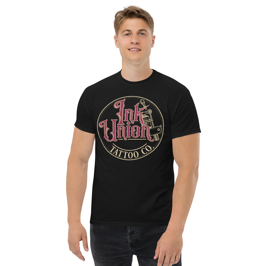 An attractive man wearing a black t-shirt with a gold circle containing fancy lettering in red and gold that says Ink Union and a gold tattoo machine peeking out from behind on the right side.  There is a dot work gradient inside the circle, and the words Tattoo Co. in gold are at the bottom of the design.
