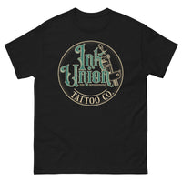 A black t-shirt with a gold circle containing fancy lettering in green and gold that says Ink Union and a gold tattoo machine peeking out from behind on the right side.  There is a dot work gradient inside the circle, and the words Tattoo Co. in gold are at the bottom of the design.