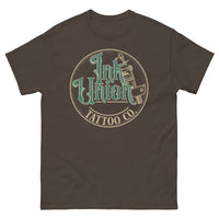 A dark brown t-shirt with a gold circle containing fancy lettering in green and gold that says Ink Union and a gold tattoo machine peeking out from behind on the right side.  There is a dot work gradient inside the circle, and the words Tattoo Co. in gold are at the bottom of the design.