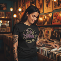 Purple & Gold Machine Tee — Tattooed Middle Eastern female model wearing black Ink Union Tattoo Co. purple and gold flash art tattoo machine graphic tee browsing records at vintage record shop with warm Edison bulb lighting — alternative streetwear, tattoo artist t-shirt, unisex graphic tee, edgy cool fashion