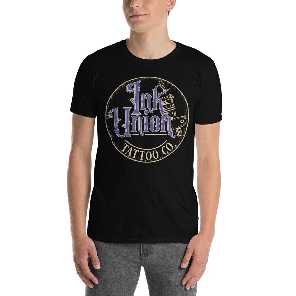 An attractive man wearing a black t-shirt with a gold circle containing fancy lettering in blue and gold that says Ink Union and a gold tattoo machine peeking out from behind on the right side.  There is a dot work gradient inside the circle, and the words Tattoo Co. in gold are at the bottom of the design.