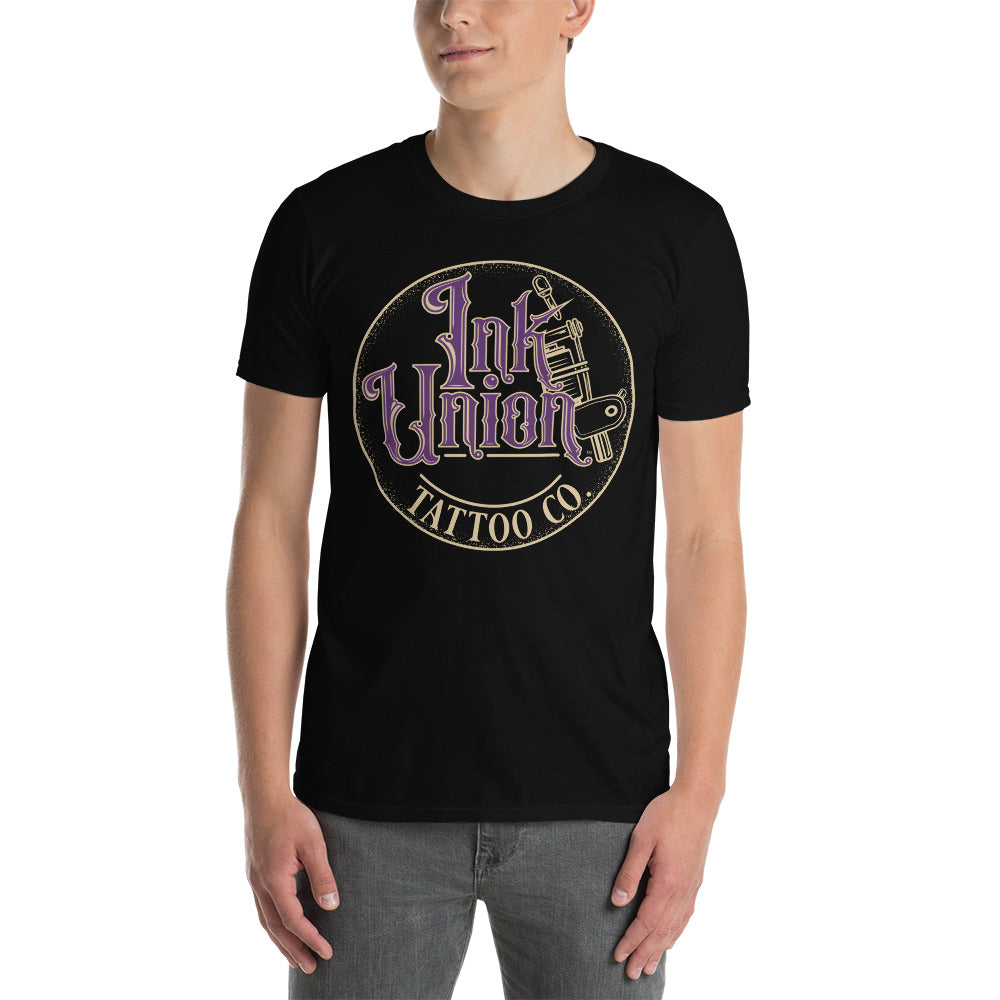 An attractive man wearing a black t-shirt with a gold circle containing fancy lettering in purple and gold that says Ink Union and a gold tattoo machine peeking out from behind on the right side.  There is a dot work gradient inside the circle, and the words Tattoo Co. in gold are at the bottom of the design.