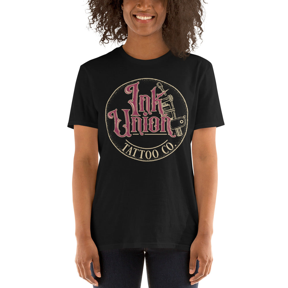 An attractive woman wearing a black t-shirt with a gold circle containing fancy lettering in red and gold that says Ink Union and a gold tattoo machine peeking out from behind on the right side.  There is a dot work gradient inside the circle, and the words Tattoo Co. in gold are at the bottom of the design.