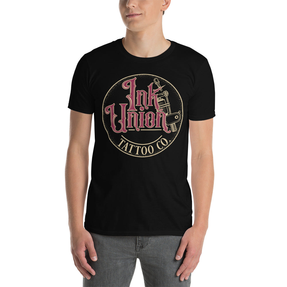 An attractive man wearing a black t-shirt with a gold circle containing fancy lettering in red and gold that says Ink Union and a gold tattoo machine peeking out from behind on the right side.  There is a dot work gradient inside the circle, and the words Tattoo Co. in gold are at the bottom of the design.