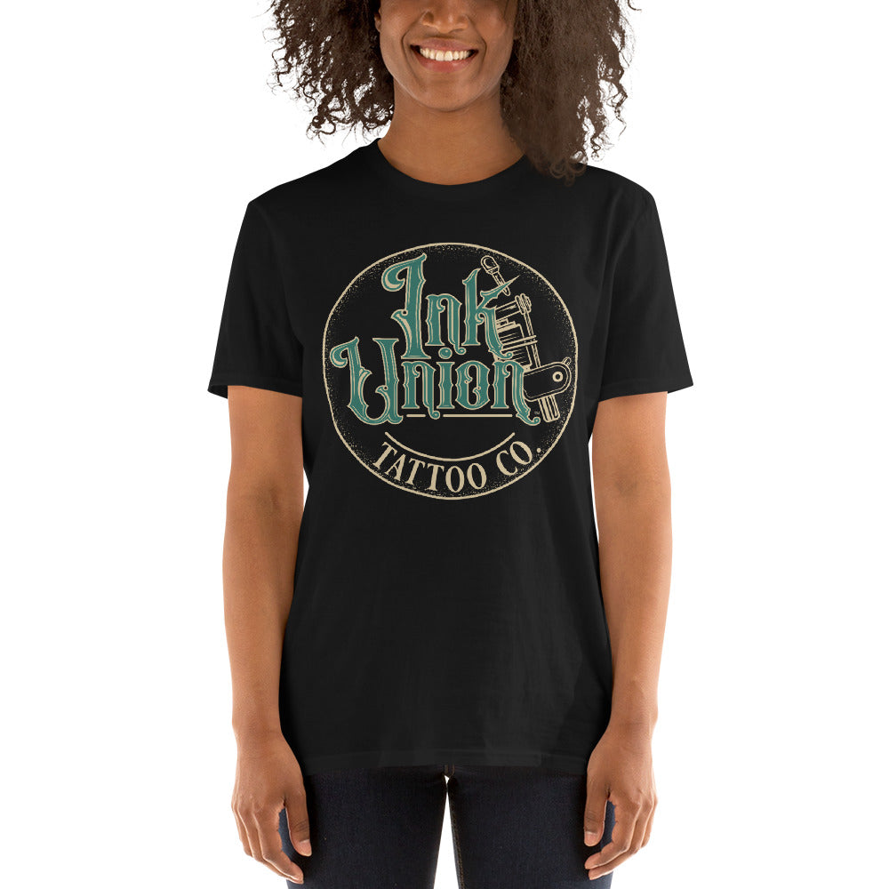 An attractive woman wearing a black t-shirt with a gold circle containing fancy lettering in green and gold that says Ink Union and a gold tattoo machine peeking out from behind on the right side.  There is a dot work gradient inside the circle, and the words Tattoo Co. in gold are at the bottom of the design.