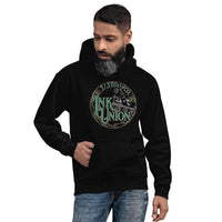 An attractive man wearing a black hoodie with a gold circle containing fancy lettering in green and gold that says Ink Union and a gold tattoo machine peeking out from behind on the right side.  There is a dot work gradient inside the circle, and the words Tattoo Co. in gold are at the bottom of the design.