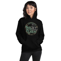 An attractive woman wearing a black hoodie with a gold circle containing fancy lettering in green and gold that says Ink Union and a gold tattoo machine peeking out from behind on the right side.  There is a dot work gradient inside the circle, and the words Tattoo Co. in gold are at the bottom of the design.
