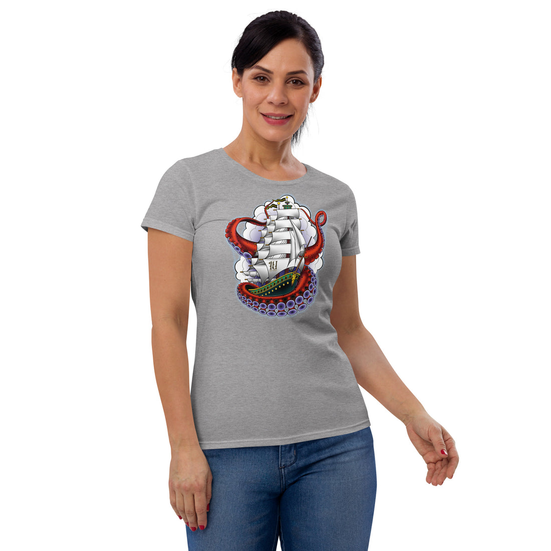 A woman wearing a grey T-shirt.  The shirt has an old-school clipper ship tattoo design in green and brown with white sails surrounded by octopus tentacles in shades of red with purple tentacles. Behind the ship are purple-tinged clouds.