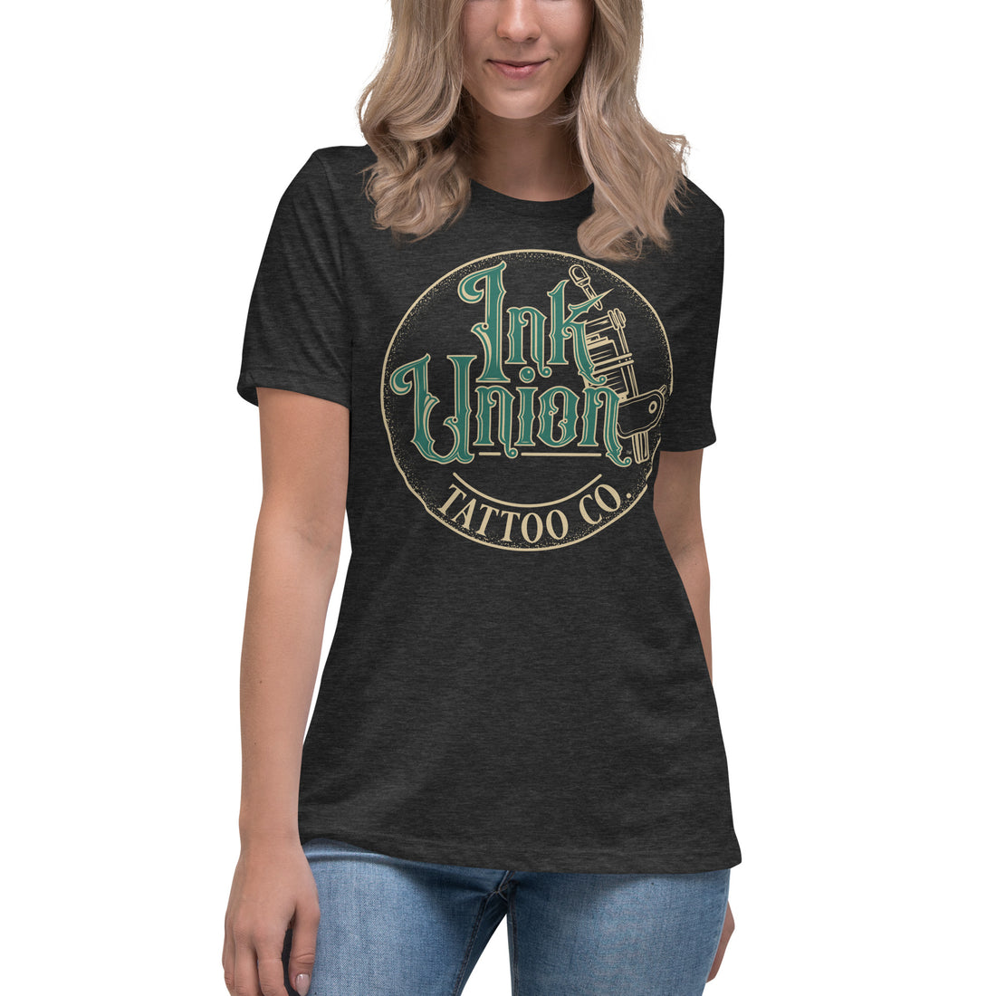 An attractive woman wearing a dark grey t-shirt with a gold circle containing fancy lettering in green and gold that says Ink Union and a gold tattoo machine peeking out from behind on the right side.  There is a dot work gradient inside the circle, and the words Tattoo Co. in gold are at the bottom of the design.