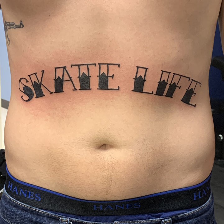 A mans stomach with with words skate life tattooed in a traditional tattoo font.