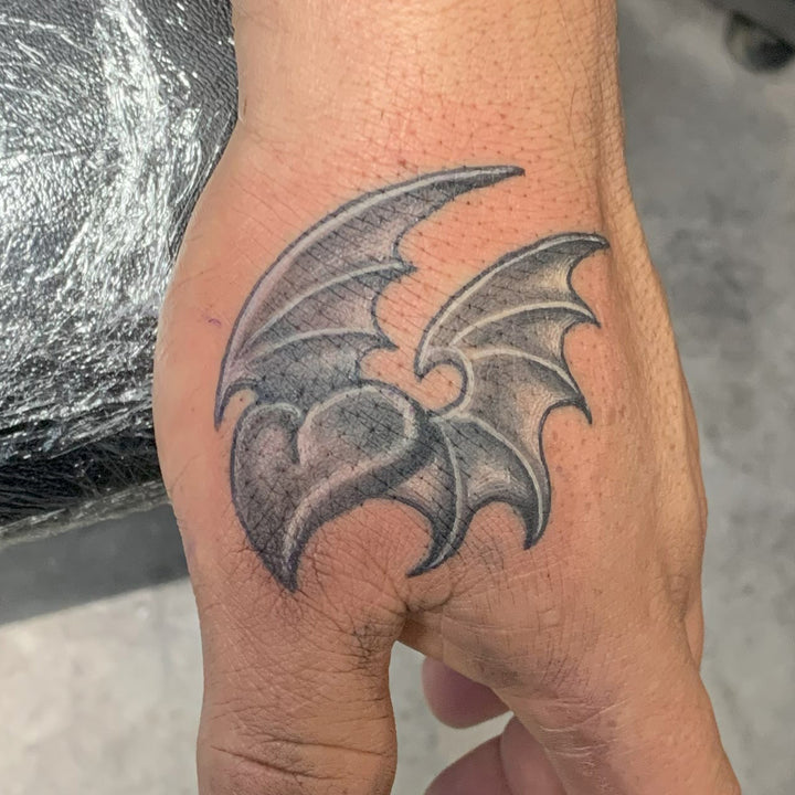 a heart with bat wings tattoo in black and grey between the thumb and wrist on a mans hand.