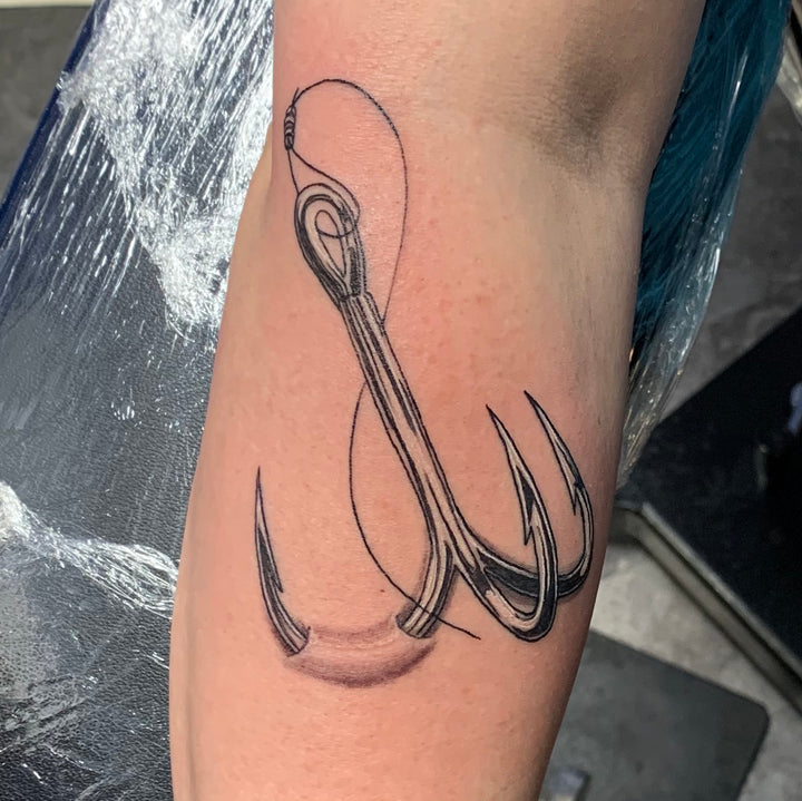 A realistic black and grey tattoo of a fishing hook embedded in a woman's forearm
