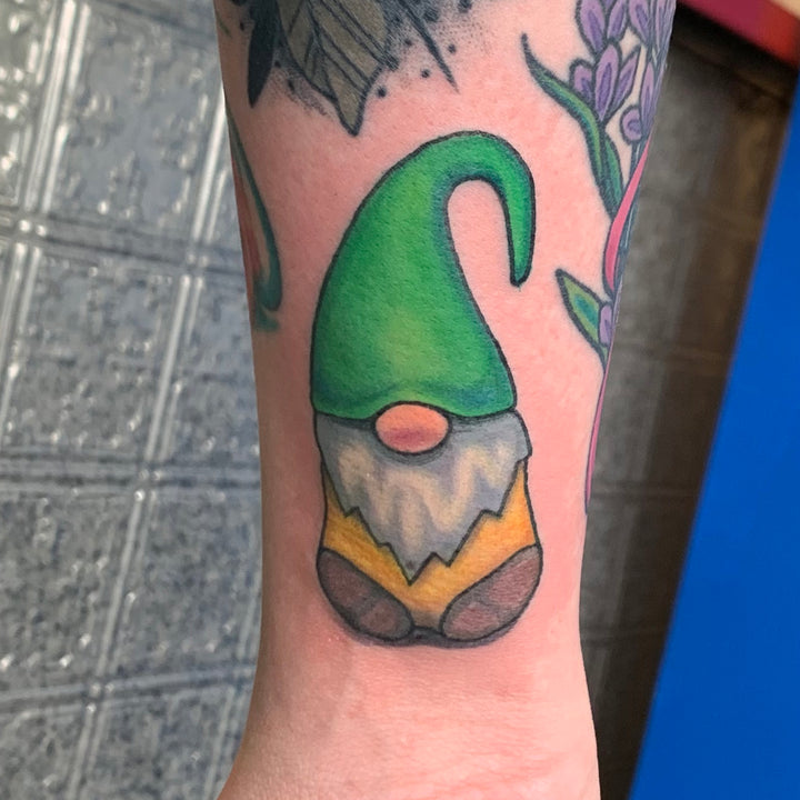 Color tattoo of a small knome with a green hat, a large nose, white beard, and a yellow shirt.