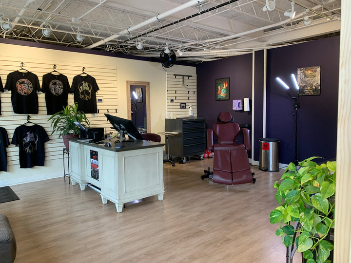A picture of Ink Union Tattoo Co. Studio featuring a clean shop with Ink Union T-shirts on  one wall , art work on the other walls. The shop has a wood floor and is painted in cream and dark purple. The photo features the new client chair also.