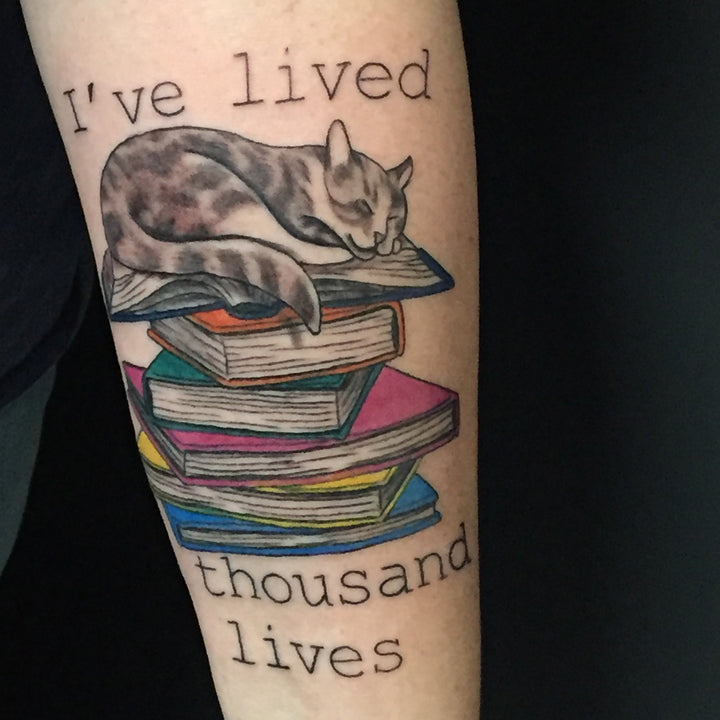 A color tattoo of a cat lying on top of a stack of books and the words I've lived a thousands lives.