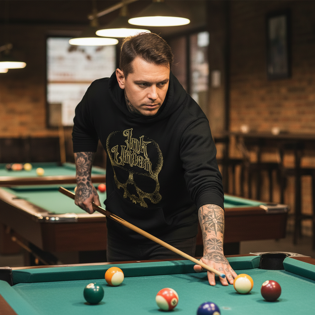 Black & Gold Dot Work Skull Hoodie — Tattooed male model wearing black Ink Union Tattoo Co. black and gold dot work skull hoodie leaning over pool table mid-shot — alternative streetwear, tattoo artist sweatshirt, unisex heavy blend hoodie, edgy cool fashion

