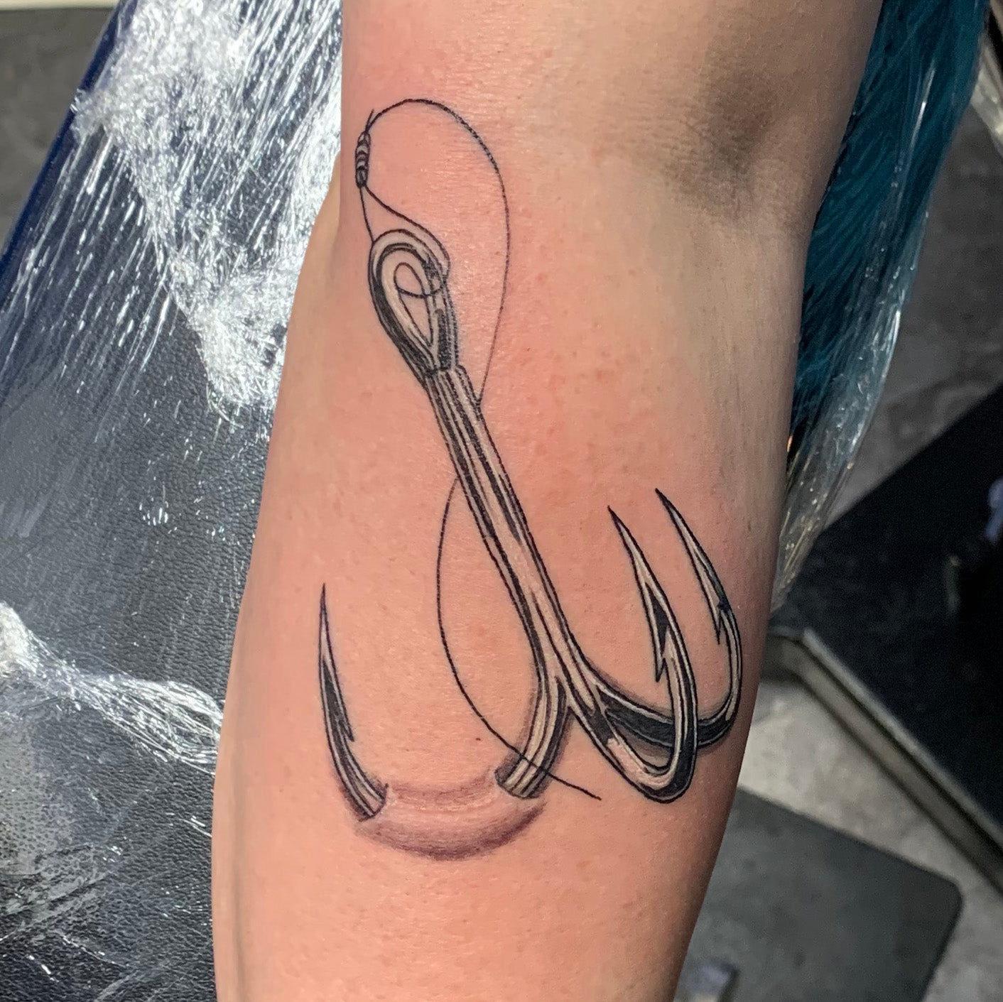 realistic black and grey tattoo of a fish hook thats embeded in the persons arm