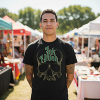 Green & Gold Dot Work Skull Tee — Tattooed Native American male model wearing black Ink Union Tattoo Co. green and gold dot work skull graphic tee at outdoor farmers market with colorful vendor tents and green trees in background — alternative streetwear, tattoo artist t-shirt, unisex graphic tee, edgy cool fashion
