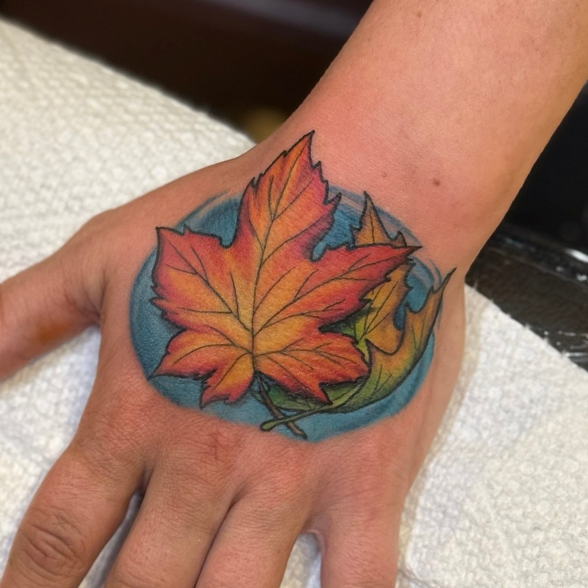 color hand tattoo of maple leaves twisting in the wind with a blue backgroumd