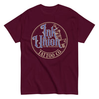 A maroon t-shirt with a gold circle containing fancy lettering in blue and gold that says Ink Union and a gold tattoo machine peeking out from behind on the right side.  There is a dot work gradient inside the circle, and the words Tattoo Co. in gold are at the bottom of the design.