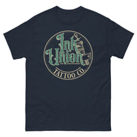 A navy blue t-shirt with a gold circle containing fancy lettering in green and gold that says Ink Union and a gold tattoo machine peeking out from behind on the right side.  There is a dot work gradient inside the circle, and the words Tattoo Co. in gold are at the bottom of the design.