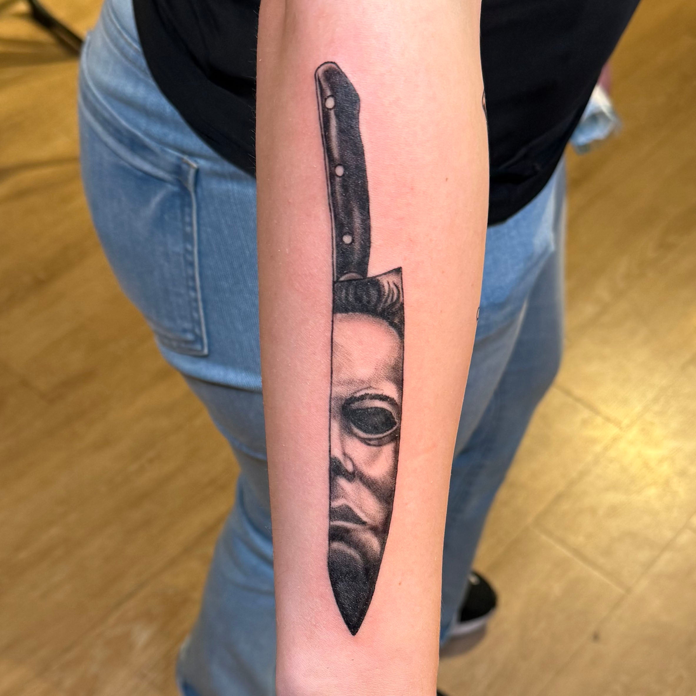 black and grey arm tattoo of a knife with the reflection of Mike Meyers, the villan of the Halloween movies, in the blade of the knife.