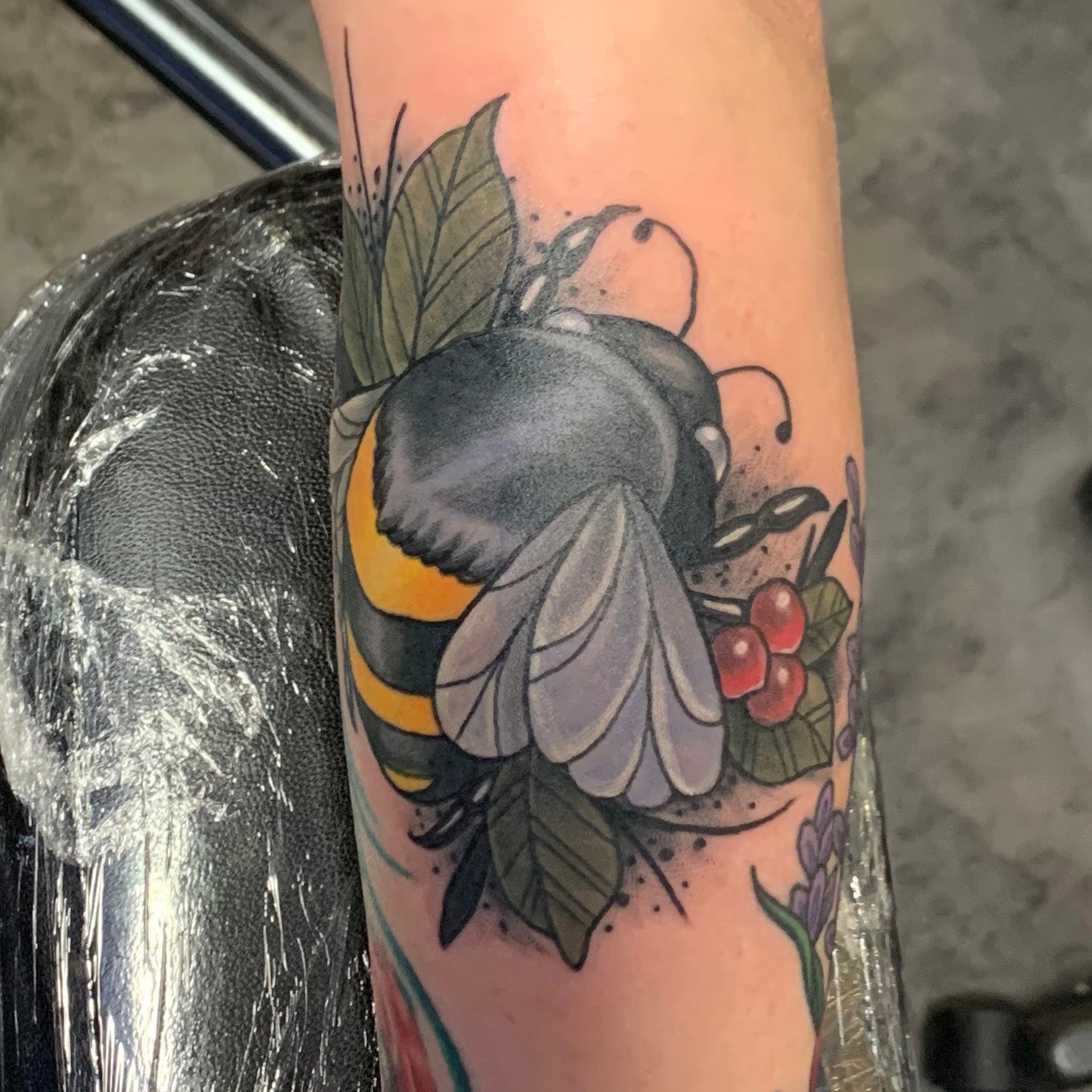 Tattoo of a bee and floral elements on a person's arm with a blurred background.