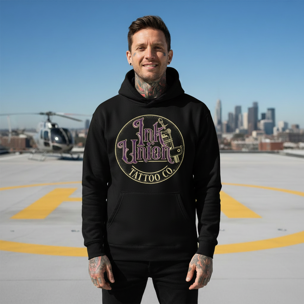 Purple & Gold Flash Art Hoodie — Tattooed male model wearing black Ink Union Tattoo Co. purple and gold flash art tattoo machine hoodie on rooftop helipad with helicopter in background — alternative streetwear, tattoo artist sweatshirt, unisex heavy blend hoodie, edgy cool fashion

