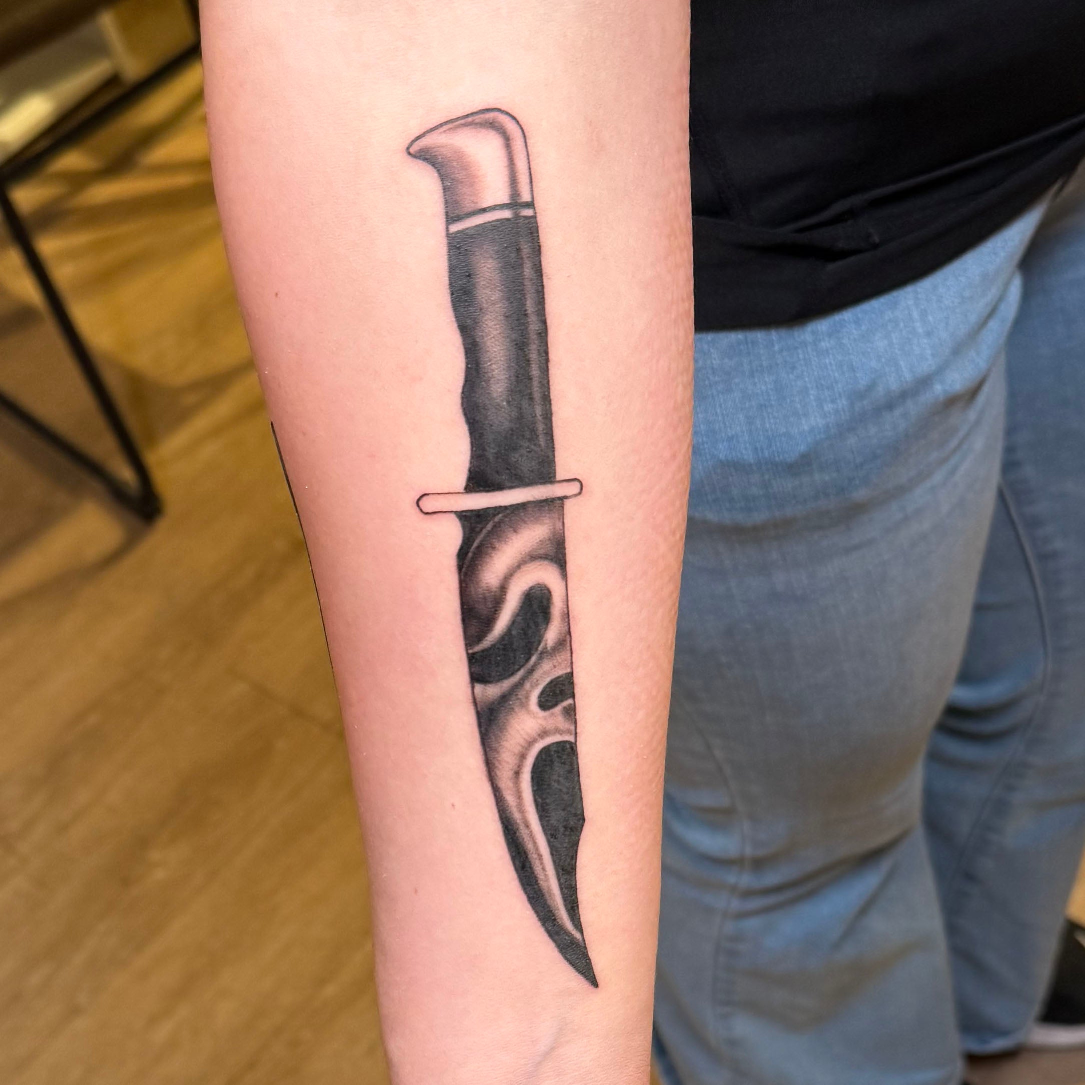 balck and grey forearm tattoo of a knife with the reflection of the ghost character from the scream movie