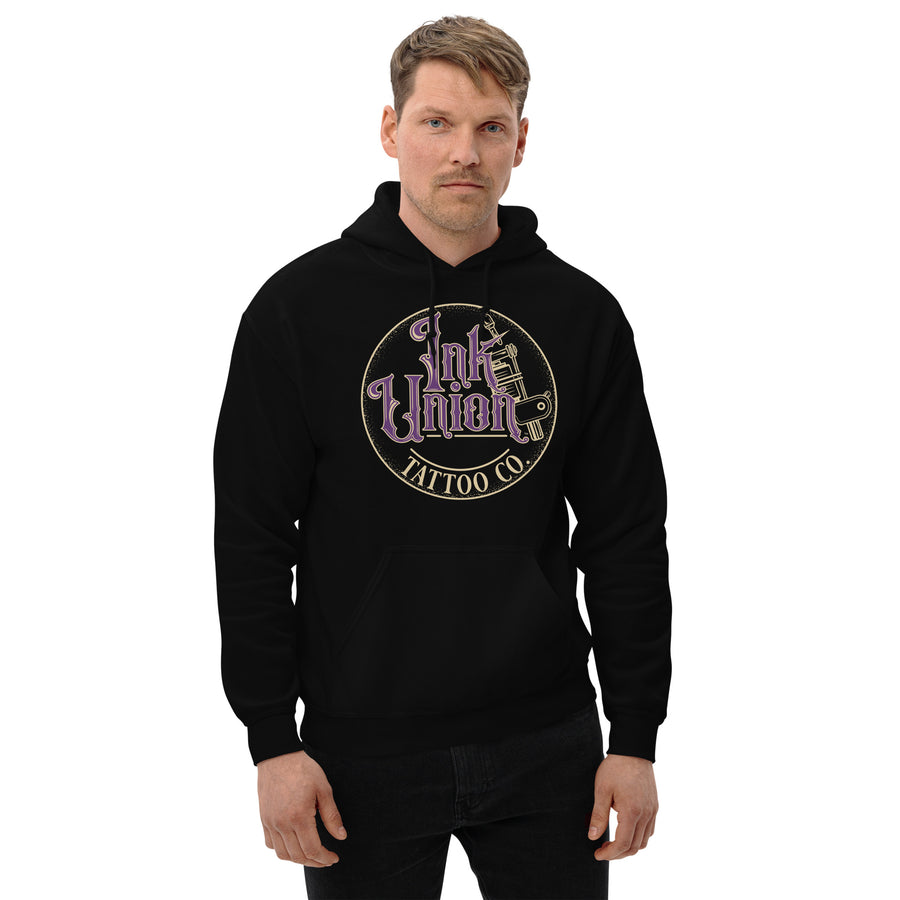 An attractive man wearing a black hoodie with a gold circle containing fancy lettering in purple and gold that says Ink Union and a gold tattoo machine peeking out from behind on the right side.  There is a dot work gradient inside the circle, and the words Tattoo Co. in gold are at the bottom of the design.