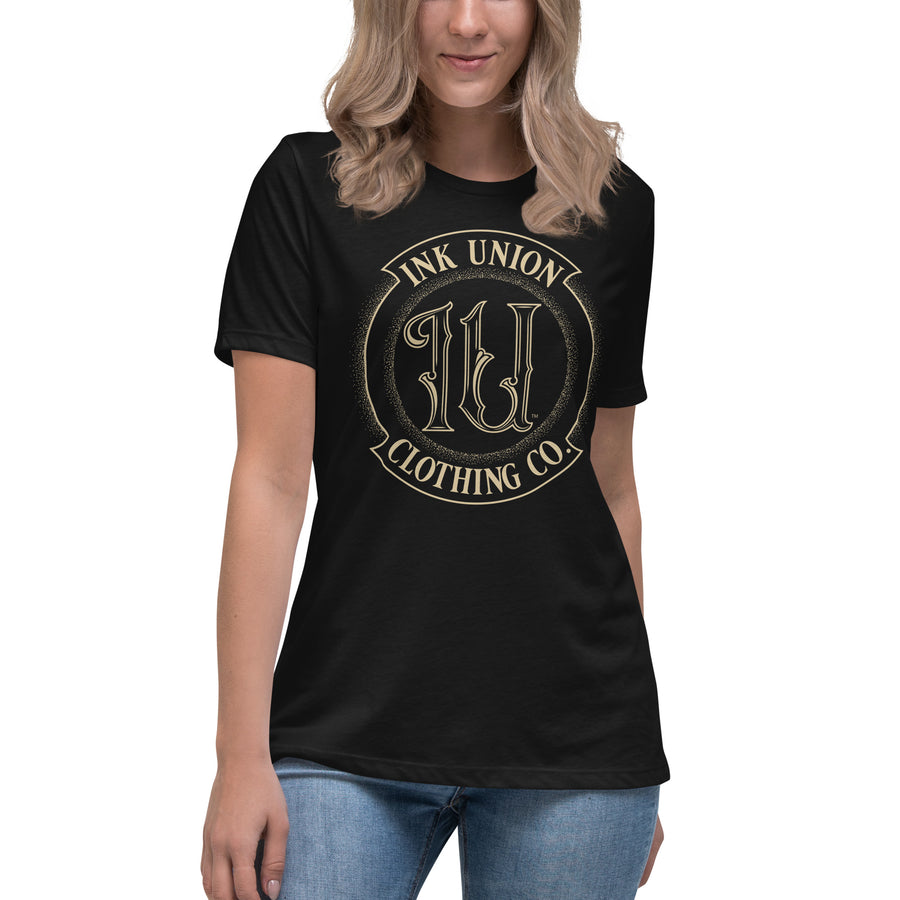 An attractive woman is wearing a black t-shirt adorned with the Ink Union Clothing Co gold badge logo containing fancy lettering and dot work gradients.