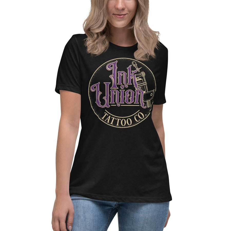 An attractive woman wearing a black t-shirt with a gold circle containing fancy lettering in purple and gold that says Ink Union and a gold tattoo machine peeking out from behind on the right side.  There is a dot work gradient inside the circle, and the words Tattoo Co. in gold are at the bottom of the design.